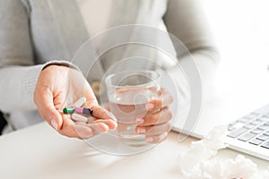 Young business woman sick in the office, hand holding pills and water glass. pills, tissue and laptop on table.