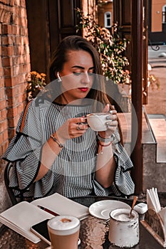 Young business woman sitting in cafe drinking coffee. Beautiful girl is working against an orange brick wall. View of freelancer