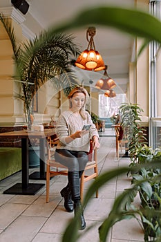 Young business woman sitting in cafe at table, leaning his hand back in chair, looking out window and holding smartphone