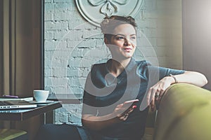 Young business woman sitting in cafe at table, leaning his hand back in chair,looking out window and holding smartphone.