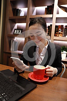 Young business woman sitting in coffee shop at wooden table, drinking coffee and using smartphone.On table is laptop