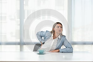 Young business woman sitting in the office in formal wear at empty desk with cup of coffee have a back pain from long stressful