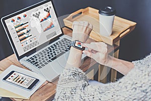 Young business woman sitting in office at table and synchronize smart watch with laptop.