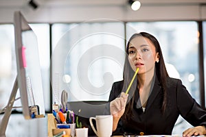 Young business woman sitting in the office is thinking about idea in front of a laptop computer looking up and  and holding pencil