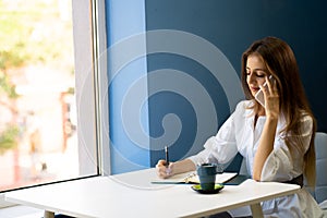 Young business woman sitting at table in cafe, talking on cell phone while taking notes in notebook with cup of coffee