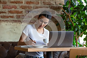 Young business woman sitting at table and taking notes in notebook.On table is laptop, smartphone and cup of coffee.On computer