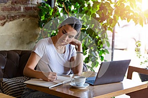 Young business woman sitting at table and taking notes in notebook.On table is laptop, smartphone and cup of coffee.On computer