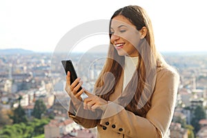 Young business woman with sweater and coat typing on smart phone outdoor with cityscape on the background
