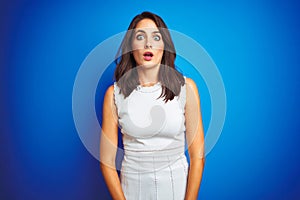 Young business woman wearing white elegant dress standing over blue isolated background afraid and shocked with surprise