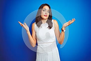 Young business woman wearing white elegant dress standing over blue isolated background clueless and confused expression with arms