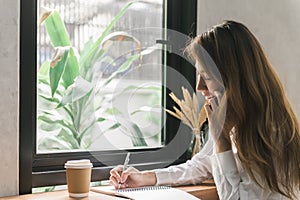 Young business woman in white dress sitting at table in cafe and writing in notebook. Asian woman talking smartphone.