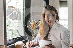 Young business woman in white dress sitting at table in cafe and writing in notebook. Asian woman using tablet.