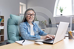 Young business woman working in office at laptop, asian smiling looking at camera, successful and happy employee