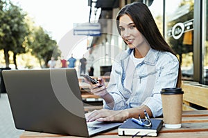 Young business woman working on open terrace at home, sitting in front of laptop, holding smartphone in her hand.