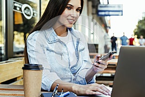 Young business woman working on open terrace at home, sitting in front of laptop, holding smartphone in her hand.