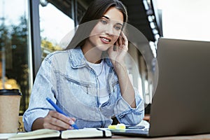 Young business woman working on open terrace at home, sitting in front of laptop, holding smartphone in her hand.