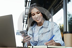 Young business woman working on open terrace home, sitting in front of laptop, taking notes in black notebook.