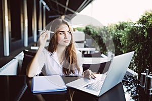 Young business woman in yellow dress sitting at table in cafe and writing in notebook on table is laptop, smartphone and cup of co