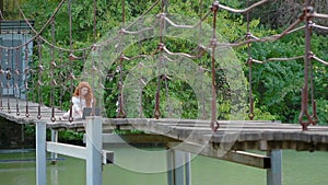 Young business women with thick beautiful fiery red hair sits on suspension bridge over river and works on laptop