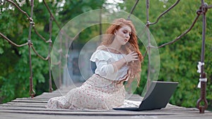 Young business women with thick beautiful fiery red hair sits on suspension bridge over river and works on laptop