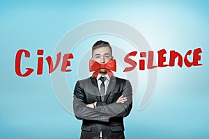A young businessman, arms folded, with a beautiful red bow covering his mouth and the title `Give Silence`.