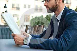 A young businessman with a beard and wearing glasses is reading a newspaper