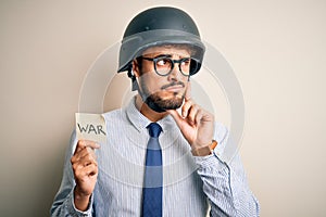 Young businessman with beard wearing helmet holding paper with war message serious face thinking about question, very confused