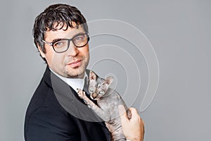 A young businessman in a black suit and glasses holds a pedigreed Sphinx cat in his hands. On a grey isolated background
