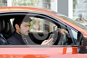 Young businessman in a black suit look at mobile phone while holding the car wheel