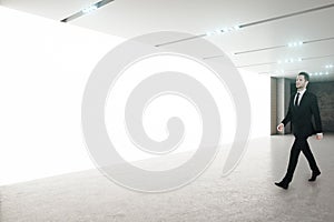 Young businessman in black suit walking in empty spacious hall room with blank white glowing wall and led lights on ceiling.