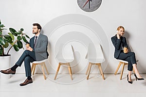 young businessman and businesswoman holding paper cups and waiting on chairs