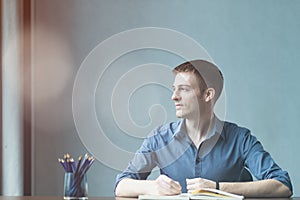 Young businessman caucasians sitting at desk office table and taking notes in notebook. Writing and looking out the window