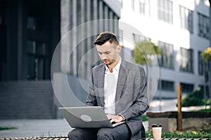 Young businessman in the city sits on bench, takes a laptop and works. Businessman typing on laptop computer outdoor