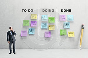 Young businessman in concrete room with creative to do lists, glued colorful paper stickers on wall. Project and schedule