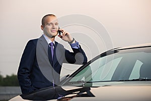 Young businessman in dark blue suit calling on cell phone leaning on his car