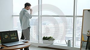 Young businessman drinking coffee looking out of window at home while laptop with business info is visible on table