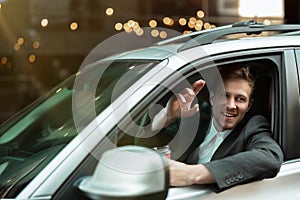 Young businessman drinks coffee and waves his friend from the open window while driving his car , multitasking, big city life