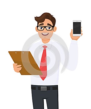 Young businessman in eye glasses holding clipboard and showing mobile, cellphone or smartphone screen. Person carrying document.