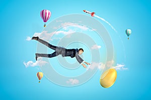 Young businessman falling down in blue sky trying to catch golden egg, with hot air balloons and rocket in background.