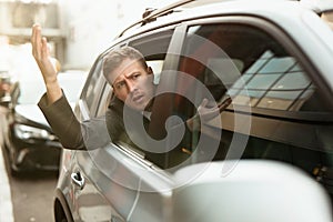 Young businessman feels nervous looking out from the open car window stuck in traffic jam, big city life