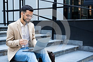 Young businessman in formal wear sitting on the stairs out of office building working on laptop computer. Job candidate work on