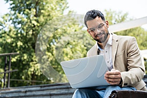 Young businessman in formal wear sitting on the stairs out of office building working on laptop computer. Job candidate work on