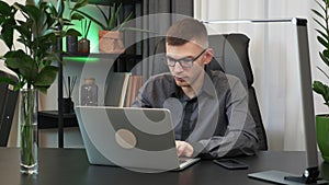 Young businessman in glasses is opening laptop computer and starting to work at modern office. Man is typing on notebook sitting i