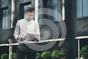 Young businessman in glasses reads documents in the street near office building