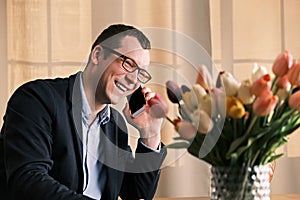 Young businessman in glasses and suit is talking with his beloved woman on mobile phone against the backdrop of bouquet