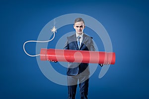 Young businessman holding big red tnt dynamite stick with lighted fuse on blue background