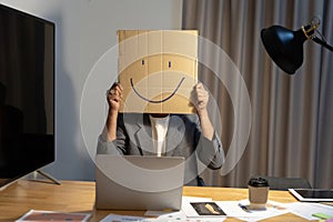 A young businessman is holding a cardboard with a smiley face on it in front of his head, gesturing with a cardboard box