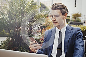 Young businessman holding mobile phone in hand and sitting next to green plant.