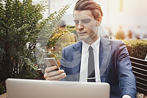 Young businessman holding mobile phone in hand and sitting next to laptop.
