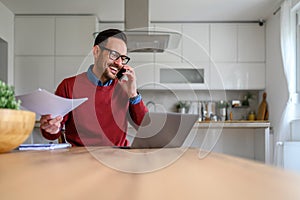 Young businessman holding tax forms and talking over mobile phone with laptop on desk in home office